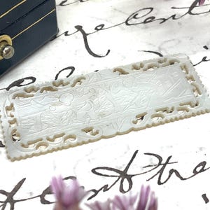 May include: Two rectangular, off-white carved mother-of-pearl plaques with intricate relief scenes of figures and buildings. The plaques have a decorative, openwork border and are set against a patterned background. A black box is in the background.