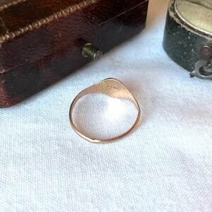 May include: A gold ring with a rounded triangular face. The ring is set against a white fabric background, with a brown jewelry box visible in the background. The ring has a smooth, polished finish.
