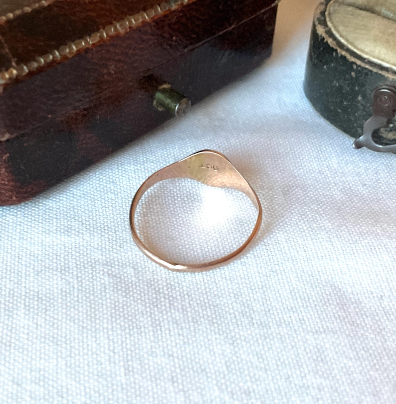 May include: A gold ring with a rounded triangular face. The ring is set against a white fabric background, with a brown jewelry box visible in the background. The ring has a smooth, polished finish.