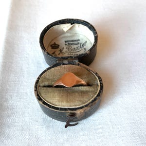 May include: A rose gold signet ring with a smooth, oval face rests inside an open, antique black leather ring box. The box is lined with green velvet and has a clasp. The box lid has text that reads "Watchmaker & Jeweller A.H. Baxler 5 Mark".
