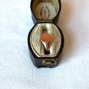 May include: A rose gold signet ring with an oval face, displayed inside a vintage brown leather jewelry box. The box is lined with cream-colored fabric and has a hinged lid. The ring is a classic piece of jewelry.