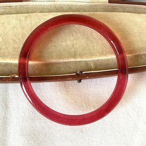 May include: A translucent, red bangle bracelet. The circular bracelet is made of a solid material and is resting on a light-colored surface. The bracelet is a simple, elegant piece of jewelry.