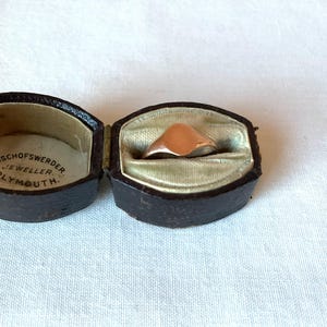 May include: A rose gold signet ring displayed in an open, antique, dark brown leather jewelry box with a light green satin lining. The box is embossed with the text "BISCHOFSWERDER JEWELLER PLYMOUTH."