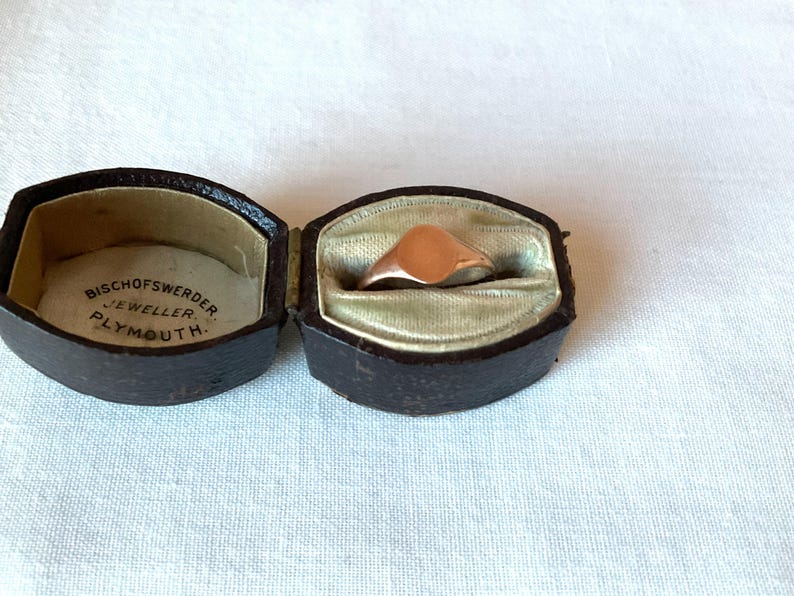 May include: A rose gold signet ring displayed in an open, antique, dark brown leather jewelry box with a light green satin lining. The box is embossed with the text "BISCHOFSWERDER JEWELLER PLYMOUTH."
