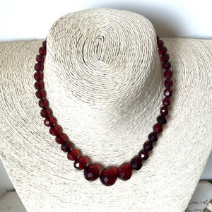 May include: A red glass bead necklace with a graduated design. The necklace is made of faceted beads and has a center cluster of larger beads.