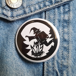 Könnte beinhalten: Ein schwarz-weißer Knopf mit einer Silhouette einer Hexe, die über einem Lagerfeuer fliegt. Die Hexe trägt einen spitzen Hut und ein langes, fließendes Gewand. Das Lagerfeuer besteht aus Holzscheiten und hat Flammen, die aus ihm herauskommen.