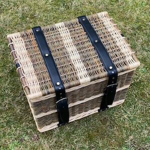 May include: A woven wicker basket with a light brown and white pattern. The basket has two black leather straps with brass buckles.
