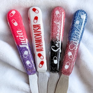 May include: Four nail art tools with glitter handles in pink, white, black, and blue. Each tool has a different name written on it: Lucia, Haykoush, Savannah, and Sofia. The handles are decorated with pill-shaped designs.