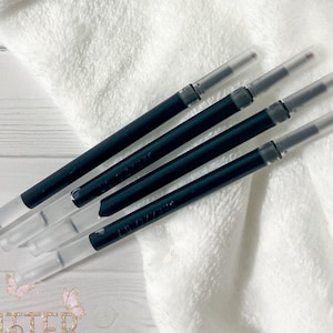 May include: Four black pen refills with clear tips are arranged diagonally on a white, textured surface. The pen refills have a sleek design, with a dark barrel and a transparent tip. The image also includes the text "Glitter Babes" with butterfly graphics.