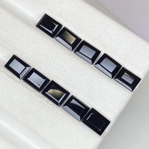 Puede incluir: Dos filas de gemas negras rectangulares. Cada gema tiene una superficie pulida y reflectante. Las gemas se muestran sobre una superficie blanca y texturizada, creando un contraste entre las piedras oscuras y el fondo claro.