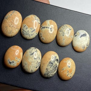 May include: Nine oval gemstones in a collection. The stones have a beige base colour with orange and black speckles. The polished stones are smooth and reflect light.