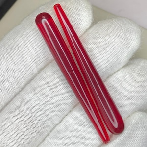 Pode incluir: Duas varas longas e finas de vidro vermelho translúcido. Uma vara tem uma extremidade arredondada com um pequeno orifício, enquanto a outra é pontiaguda. As varas estão sobre uma superfície branca e texturizada.