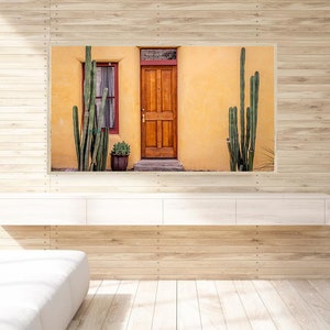 May include: A photograph of a yellow building with a wooden door and a window. There are tall green cacti growing in front of the building.