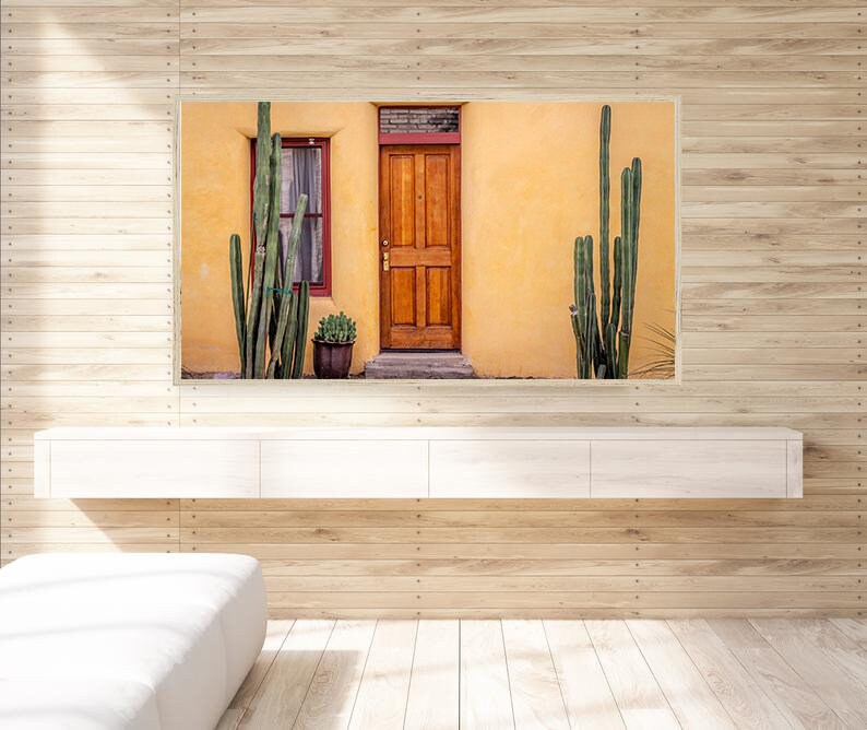 May include: A photograph of a yellow building with a wooden door and a window. There are tall green cacti growing in front of the building.
