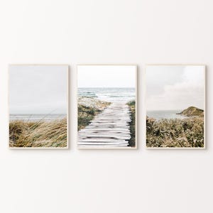 May include: A set of three framed prints depicting coastal scenes. The images show a wooden boardwalk, ocean views, and grassy dunes. The frames are light wood, creating a neutral and calming aesthetic for the home.