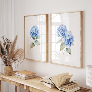 May include: Two framed watercolor prints featuring blue hydrangea flowers. The artwork has a light wooden frame and is displayed on a white wall. The prints are part of a home decor collection.