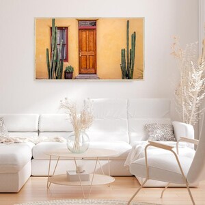 May include: A photograph of a yellow building with a wooden door and two cactus plants in front of it. The building has a window with a red frame and a small potted plant in front of it.