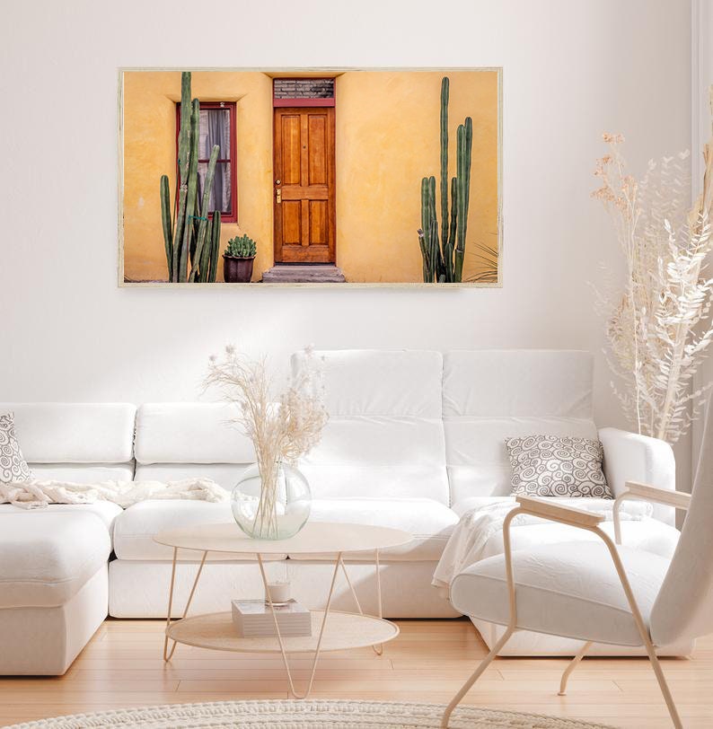 May include: A photograph of a yellow building with a wooden door and two cactus plants in front of it. The building has a window with a red frame and a small potted plant in front of it.