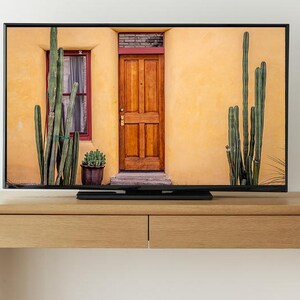 May include: A flat screen television with a black frame is mounted on a light brown wooden entertainment center. The television is displaying a picture of a yellow building with a wooden door and a window. There are tall green cacti on either side of the building.