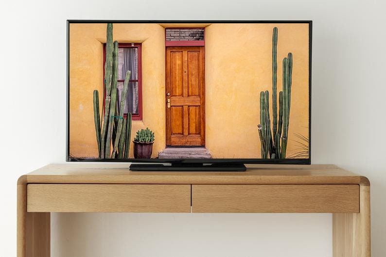 May include: A flat screen television with a black frame is mounted on a light brown wooden entertainment center. The television is displaying a picture of a yellow building with a wooden door and a window. There are tall green cacti on either side of the building.