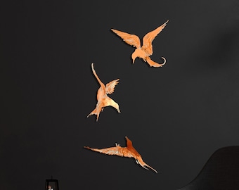 Op de afbeelding: Drie houten wandsculpturen van vogels in vlucht, met een donkergrijze muur en een zwarte stoel op de achtergrond.