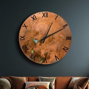 Op de afbeelding: Grote, ronde koperen wandklok met zwarte Romeinse cijfers en wijzers. De klok heeft een getextureerd oppervlak en is gemonteerd op een donkerblauwe muur boven een bruine hoekbank. De kamer is ingericht met planten en een kleine bijzettafel.