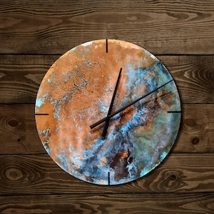 May include: Circular wall clock with a copper-effect face, displaying blue and orange abstract patterns. It has black hands and hour markers. The clock is set against a wooden background.