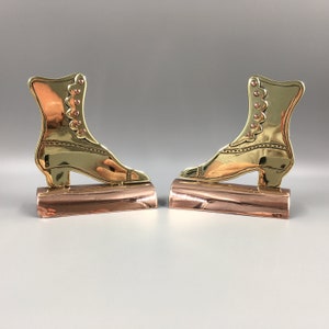 May include: A pair of brass bookends shaped like vintage boots with copper bases. The boots are gold in colour and have a detailed design.