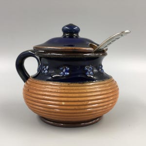 May include: A ceramic sugar bowl with a lid and handle. The bowl is dark blue with small blue floral designs and a light brown, ribbed base. A small silver spoon rests on the rim.