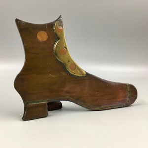 May include: A copper boot-shaped bookend with brass accents. The boot has a rounded toe and a high heel. The brass accents are in the shape of circles and a decorative design on the top of the boot.