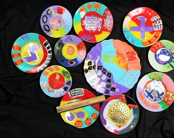 May include: A collection of 11 colorful ceramic plates with various abstract designs and patterns. The plates are painted in bright colors, including red, yellow, blue, green, purple, and orange. Some plates have geometric shapes, while others have more free-flowing designs. The plates are arranged in a circle on a black background.