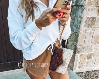 May include: A white cropped sweatshirt with a button closure and a brown,  cowhide,  tooled leather wallet with a fringe detail. The wallet is attached to a brown strap. The person is wearing brown shorts and a phone with a heart-shaped phone grip.