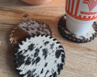 May include: Set of three round cowhide coasters with black braided edges. One coaster supports a white cup with an orange logo. The coasters have a natural, spotted pattern in black and white, and brown and white.