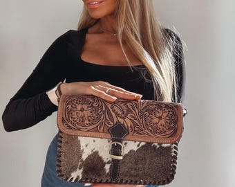 Pode incluir: Uma bolsa de ombro retangular com corpo em pele de vaca castanha e branca e aba em couro castanho escuro. A aba apresenta um intrincado padrão floral gravado e é fixada por uma tira central de couro com fecho de fivela metálica.