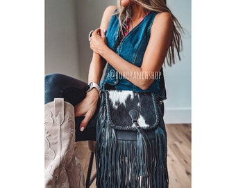 May include: A black and white cowhide crossbody bag with a fringe detail. The bag has a black strap and a silver buckle. The bag is being worn by a person wearing a denim vest and jeans. The text "@BOHORANCHSHOP" is visible in the image.