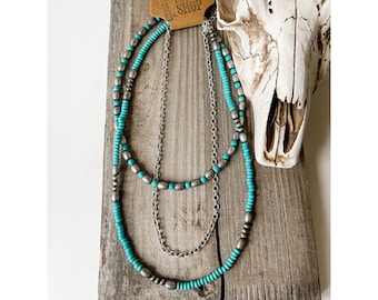 May include: A layered necklace with turquoise beads and silver chain links. The necklace is displayed on a wooden surface with a white cow skull in the background. The tag reads "Boho Ranch Shop".