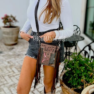May include: A clear crossbody bag with a brown leather design and fringe. The bag features a pink graphic and the text "@BOHORANCHSHOP". The person in the image is wearing a white long-sleeve shirt, denim shorts, and a white cowboy hat.