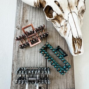 May include: Three hair claw clips in copper, silver, and teal, each with a unique beaded design. The clips are arranged on a weathered wooden surface, with a cow skull in the background. These hair accessories are perfect for adding a touch of style.
