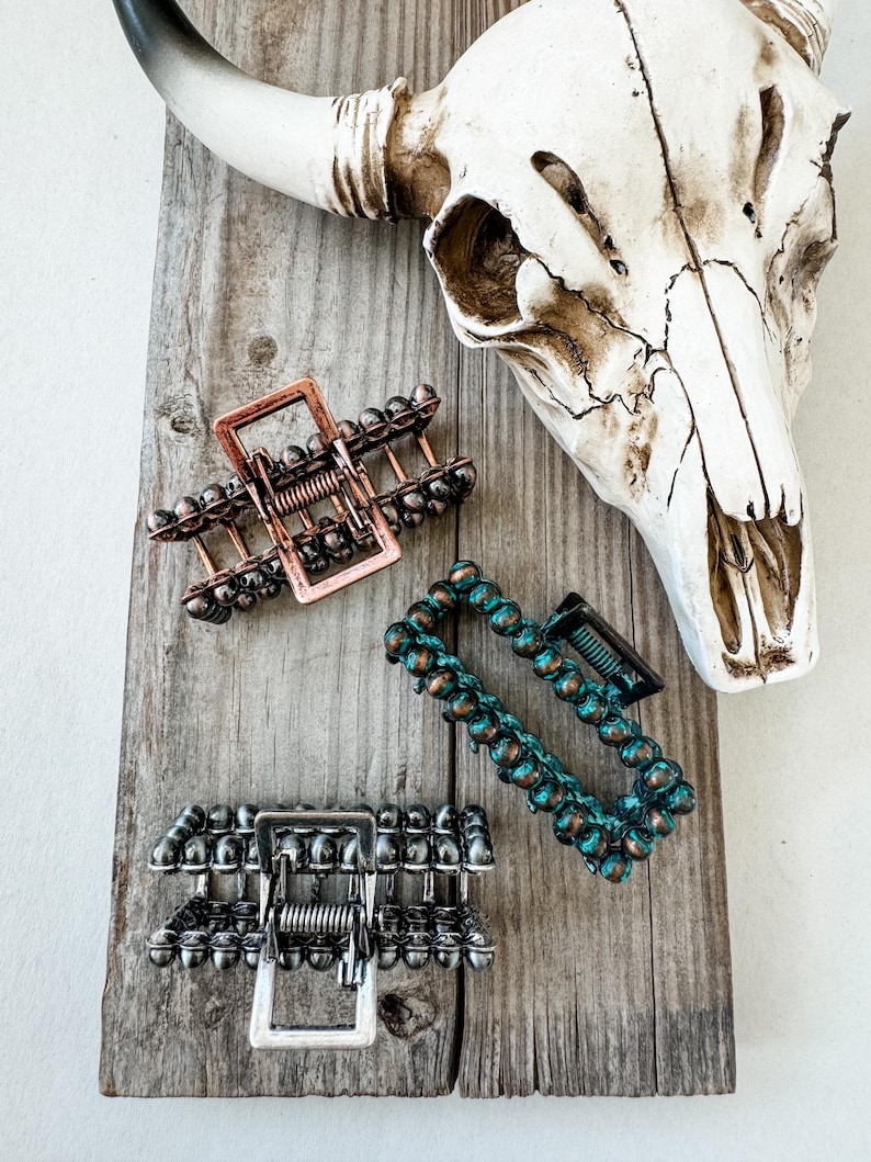 May include: Three hair claw clips in copper, silver, and teal, each with a unique beaded design. The clips are arranged on a weathered wooden surface, with a cow skull in the background. These hair accessories are perfect for adding a touch of style.