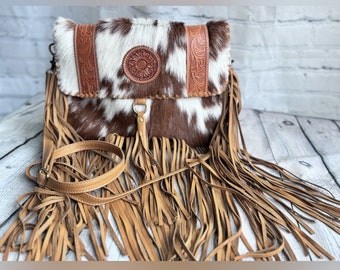 Puede incluir: Un bolso bandolera de piel de vaca marrón y blanco con ribete de flecos y una correa de cuero repujado. El bolso tiene un diseño floral en la parte delantera.