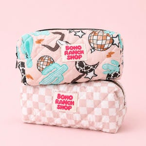 May include: Two quilted rectangular pouches stacked vertically. The top pouch is light pink with a print of disco balls, cacti, cow print moons, and stars. The bottom pouch features a light pink and white checkerboard pattern. Both have a white label with the text BOHO RANCH SHOP in pink.