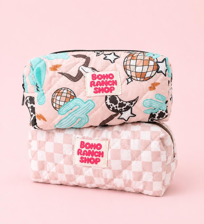 May include: Two quilted rectangular pouches stacked vertically. The top pouch is light pink with a print of disco balls, cacti, cow print moons, and stars. The bottom pouch features a light pink and white checkerboard pattern. Both have a white label with the text BOHO RANCH SHOP in pink.