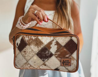 May include: A rectangular brown and tan cosmetic bag with a patchwork design. The bag features a leather handle and trim, a zipper closure, and a "Boho Ranch Shop" logo patch.