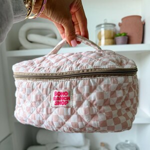 May include: A pink and white checkered quilted cosmetic bag with a brown zipper and handle. The bag has a label that reads "BOHO RANCH SHOP". The bag is held up by a hand.