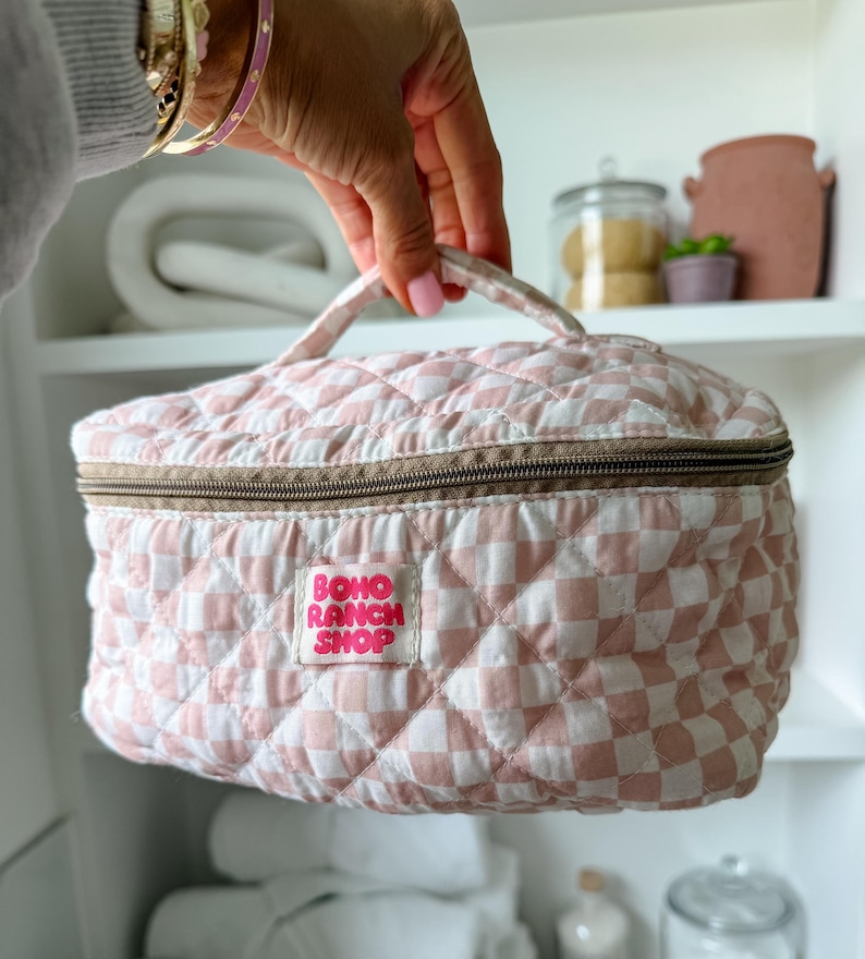 May include: A pink and white checkered quilted cosmetic bag with a brown zipper and handle. The bag has a label that reads "BOHO RANCH SHOP". The bag is held up by a hand.