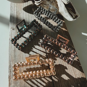 May include: Four beaded hair claw clips in gold, bronze, and gunmetal gray hues rest on a wooden surface. A cow skull is in the background. The clips are rectangular with a hinged clasp.