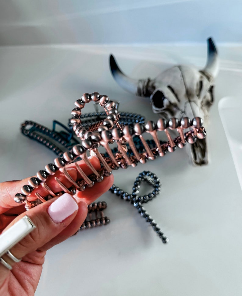 May include: A large, rose gold hair claw with a beaded design. The claw is made of metal and has a smooth, shiny finish.