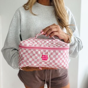 May include: A pink and white checkered cosmetic bag with a top handle and a zipper closure. The bag has a quilted design and features the text "BOHO RANCH SHOP" in a pink rectangle.