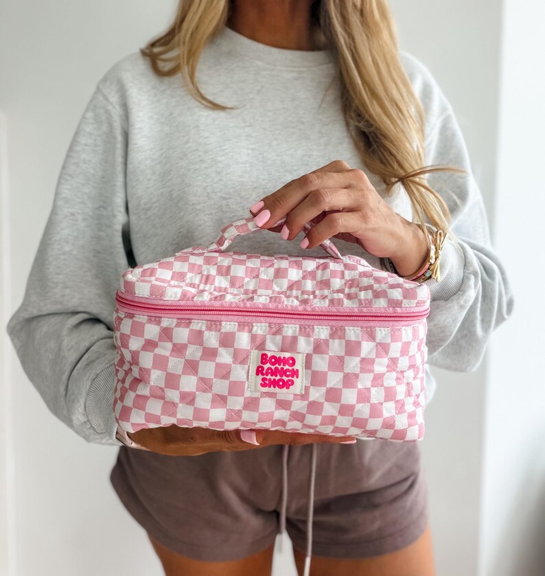 May include: A pink and white checkered cosmetic bag with a top handle and a zipper closure. The bag has a quilted design and features the text "BOHO RANCH SHOP" in a pink rectangle.
