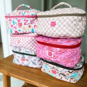 May include: A stack of quilted cosmetic bags in various patterns and colors. Designs include pink and white checkered, floral, and cowboy-themed prints. Each bag has a top handle and a zipper closure. The bags have the text "BOHO RANCH SHOP".